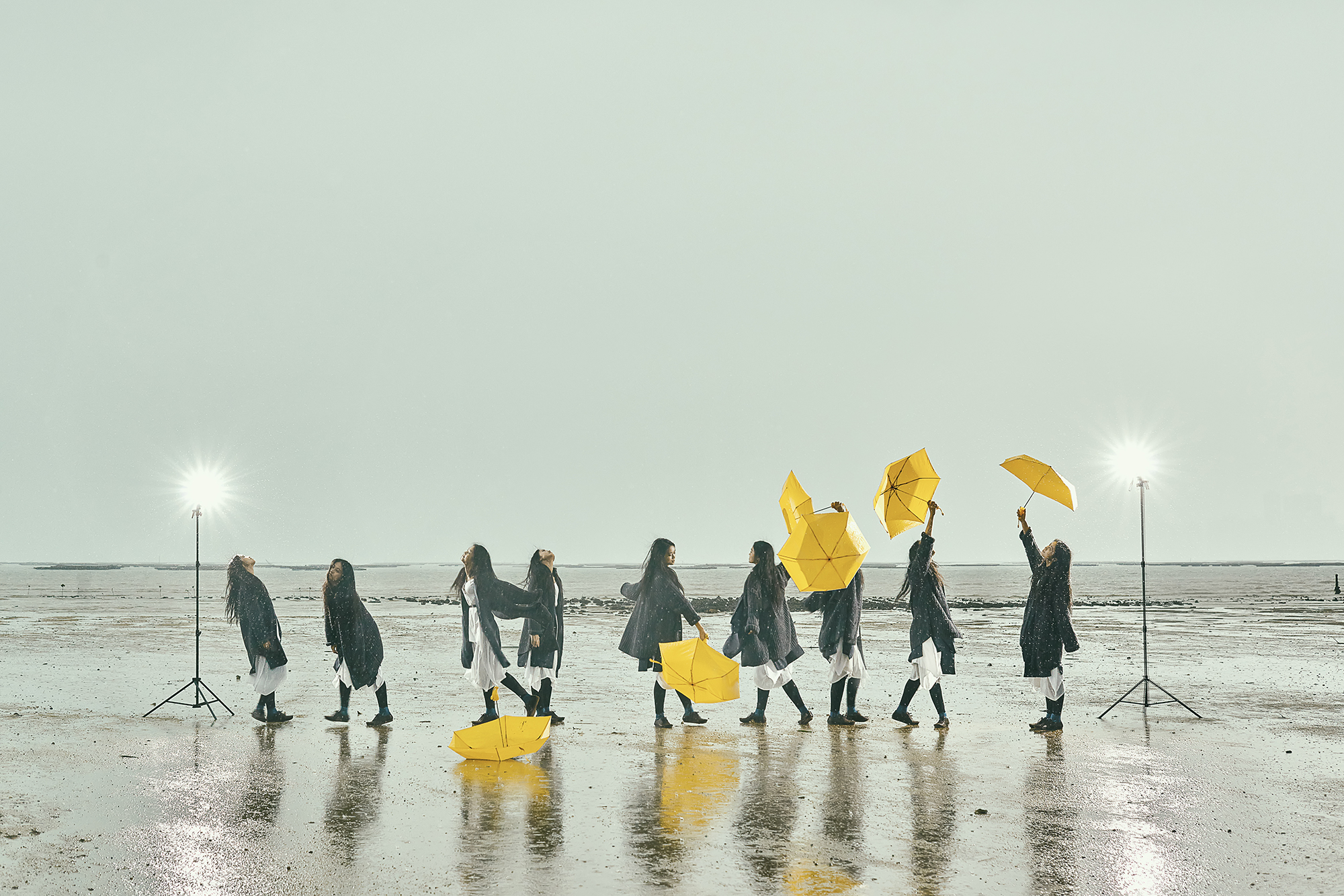 Eight people in dark coats and white outfits walk on a reflective beach, holding bright yellow umbrellas. Two lights on stands illuminate the scene.